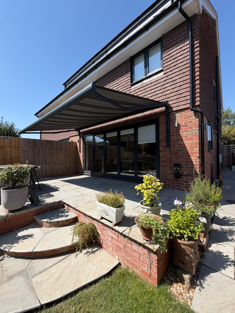 Extended anthracite awning shading a stone patio on a two-storey brick house
